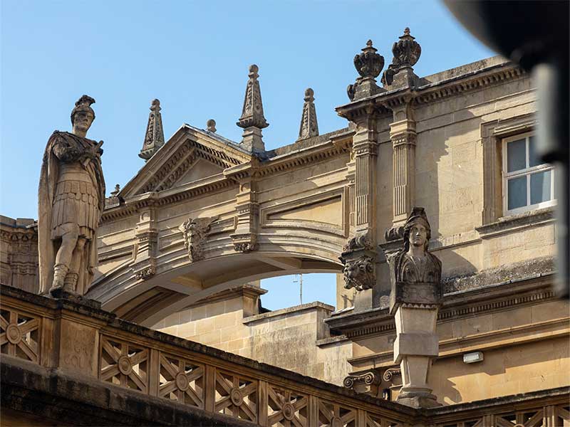 Stone architecture of the Roman Baths complex in Bath, England, featuring statues on a terrace overlooking an archway.