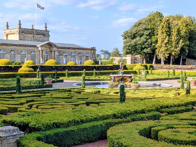 A grand view of the formal gardens at Blenheim Palace, England. UNESCO site, featuring intricate green parterre hedges surrounding a circular fountain and statue.