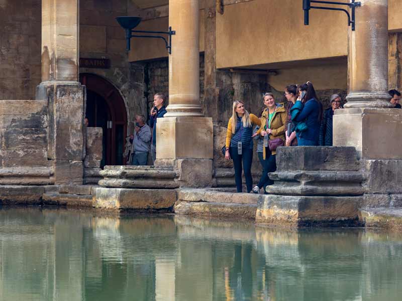 The ancient Roman Baths with green, steaming water, surrounded by historic stone columns and architecture under a modern glass roof, with several people viewing the site.