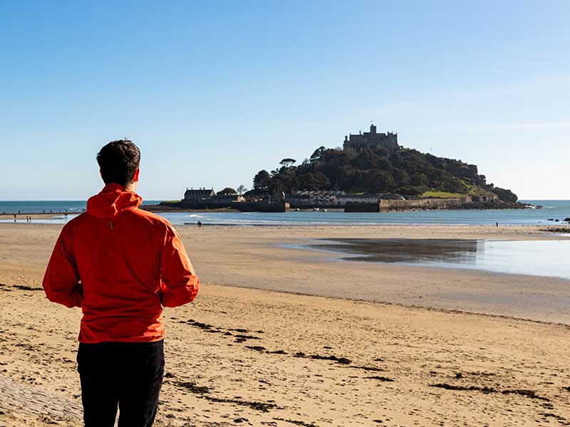 Person in an orange jacket standing on a sandy beach, looking out towards St Michael’s Mount, a historic castle-topped tidal island off the coast of Cornwall.