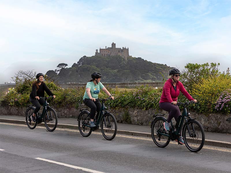 Three women on bicycles ride along a road with a prominent castle on a hill in the background.