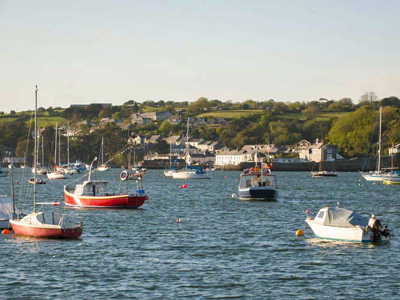A harbour dotted with colourful sailing boats and fishing vessels gently rocking in the water. Across the bay, slate-roofed houses and trees rise into the hills under soft evening light.
