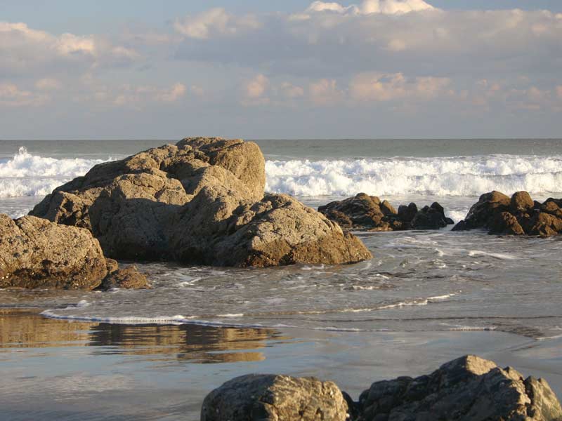 Large rocks scattered along the beach with soft evening light casting golden reflections across the wet sand and foamy waves.