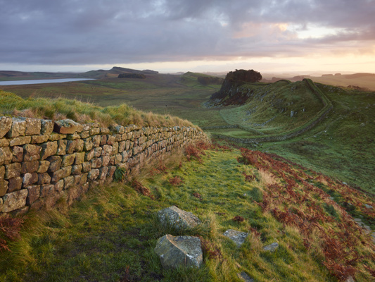 Sunrise image of Hadrian's Wall descending into a valley. The wall is covered in green moss, and the surrounding landscape is also all greenery.